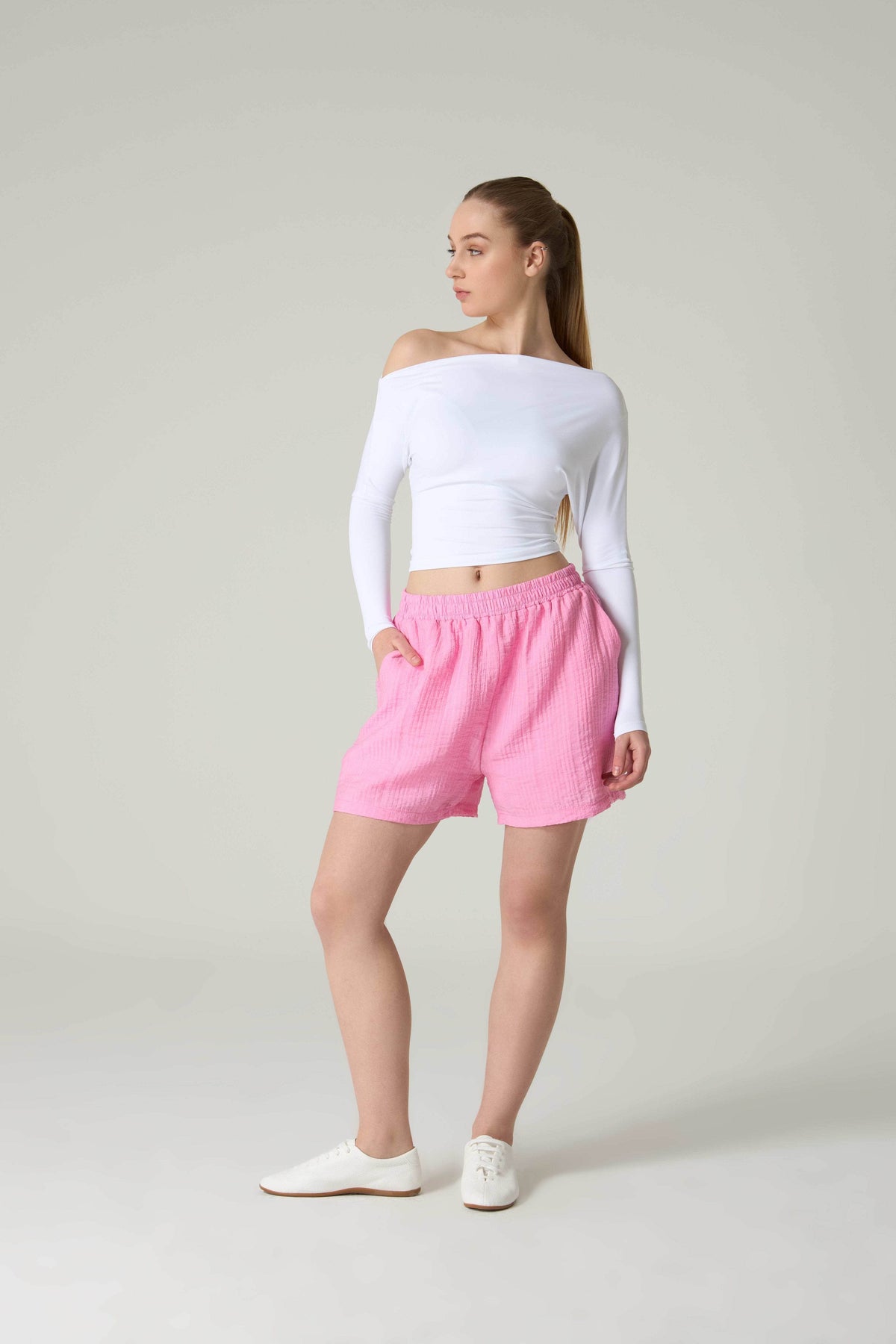 Woman wearing a white off-shoulder top and pink shorts on a plain background.