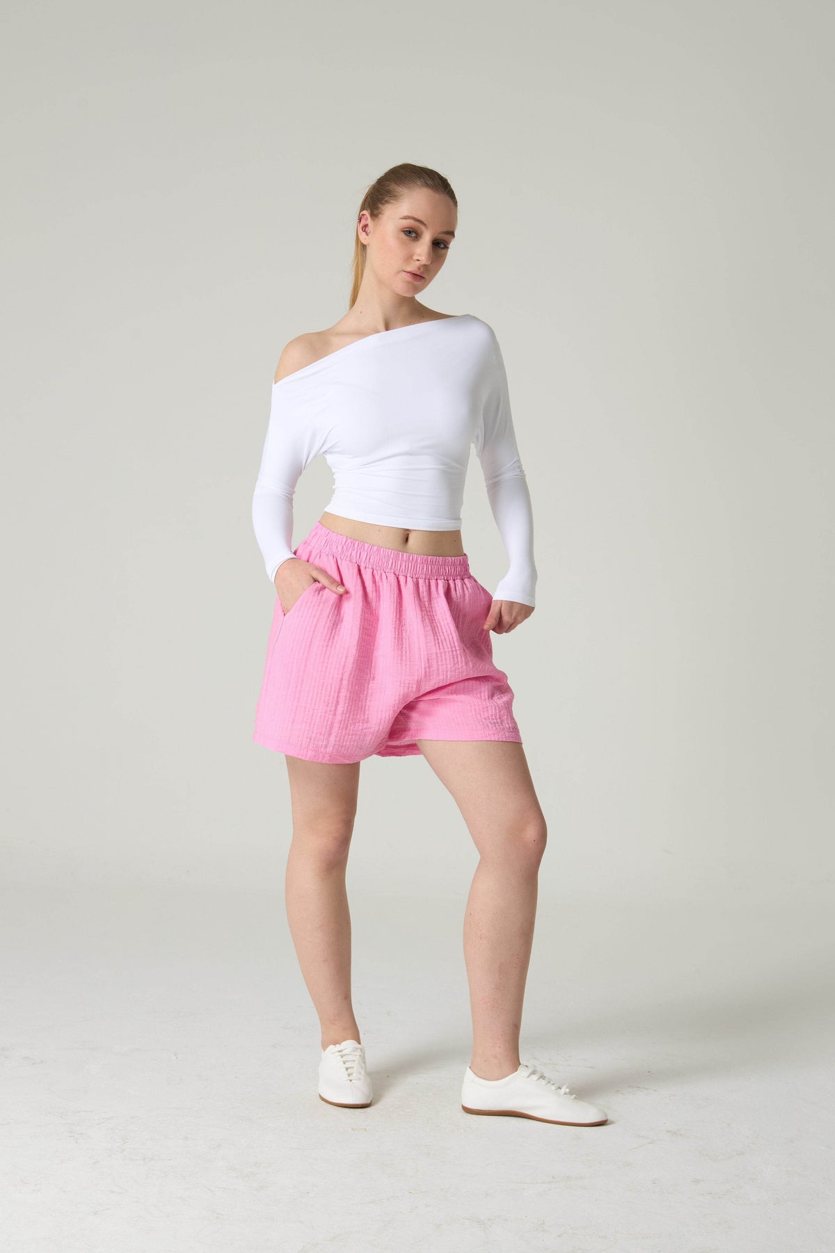Woman wearing a white off-shoulder top and pink shorts on a plain background.