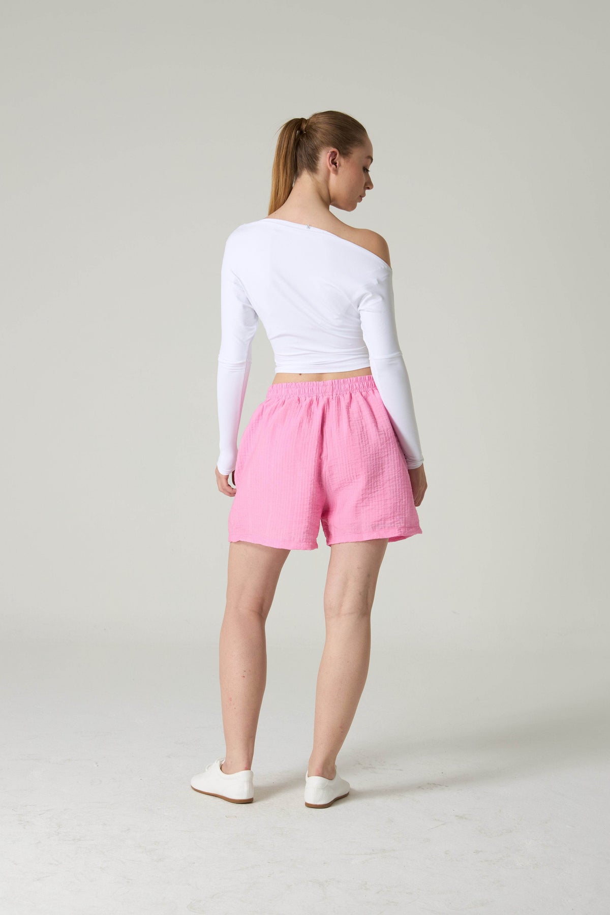 Person wearing a white off-shoulder top and pink shorts on a plain background.