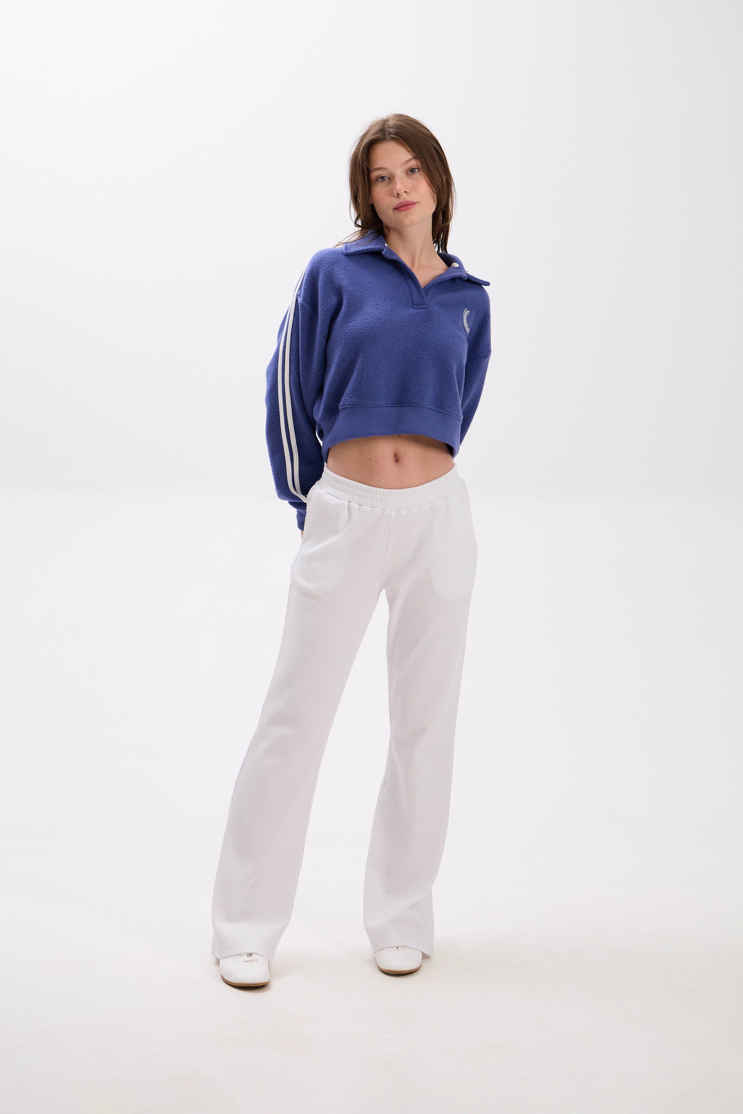 Person wearing a blue cropped sweatshirt with white stripes and white pants on a white background.