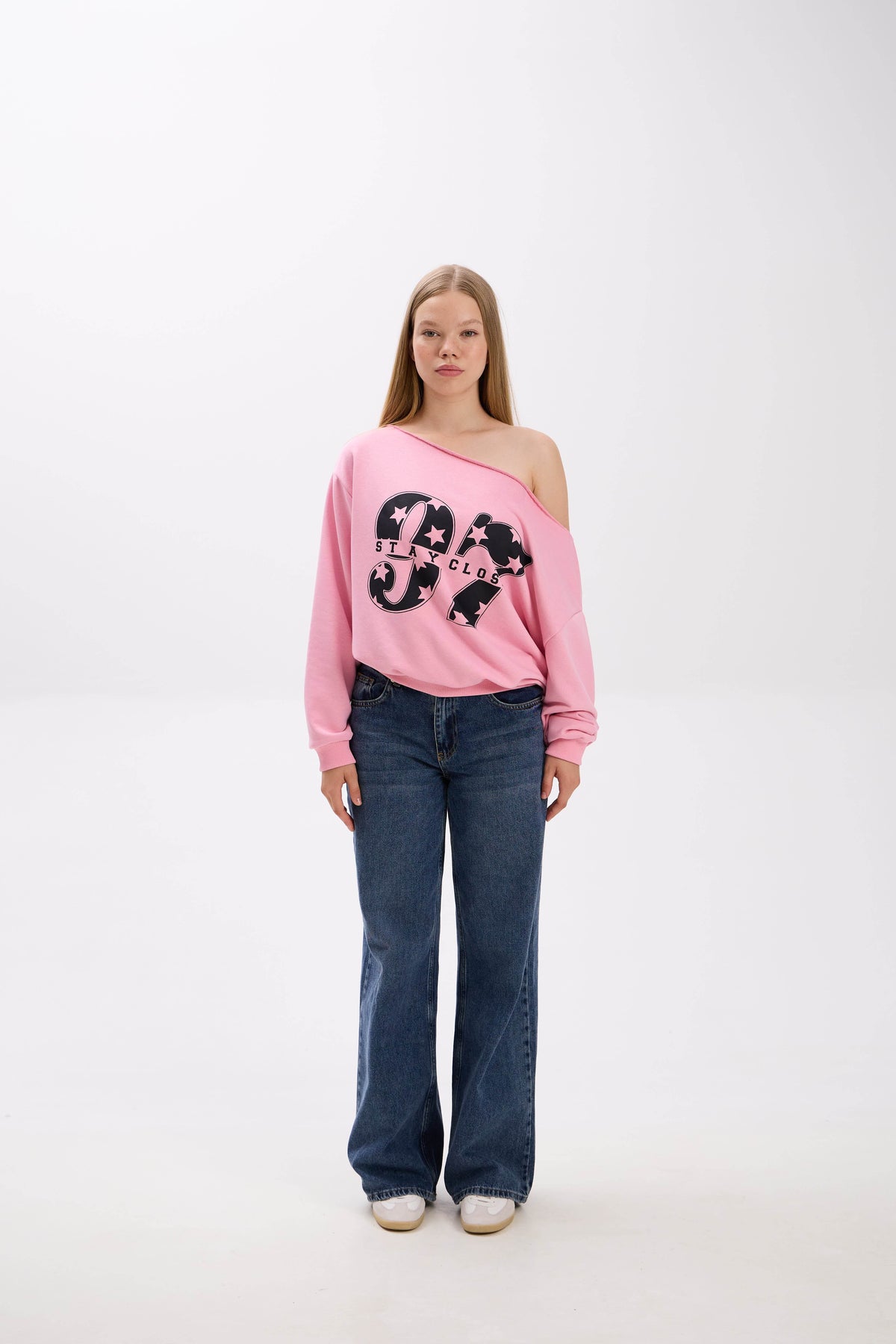 Model wearing a pink off-shoulder sweatshirt with bold “Stay Close” print, styled with blue jeans and white sneakers.
