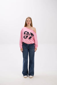 Model wearing a pink off-shoulder sweatshirt with bold “Stay Close” print, styled with blue jeans and white sneakers.