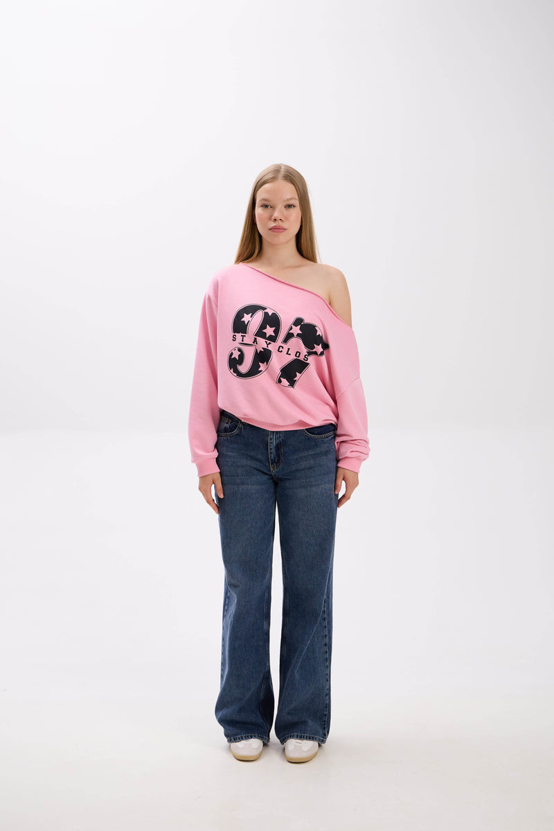 Model wearing a pink off-shoulder sweatshirt with bold “Stay Close” print, styled with blue jeans and white sneakers.