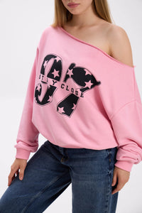 Close-up of pink off-shoulder sweatshirt with bold black “Stay Close 97” print, styled with blue jeans.