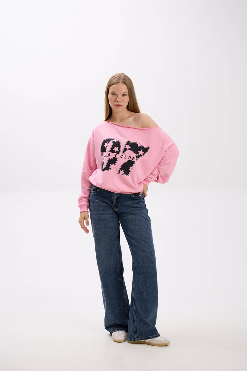 Model in a pink off-shoulder sweatshirt with bold black “Stay Close 97” print, paired with blue jeans and sneakers.