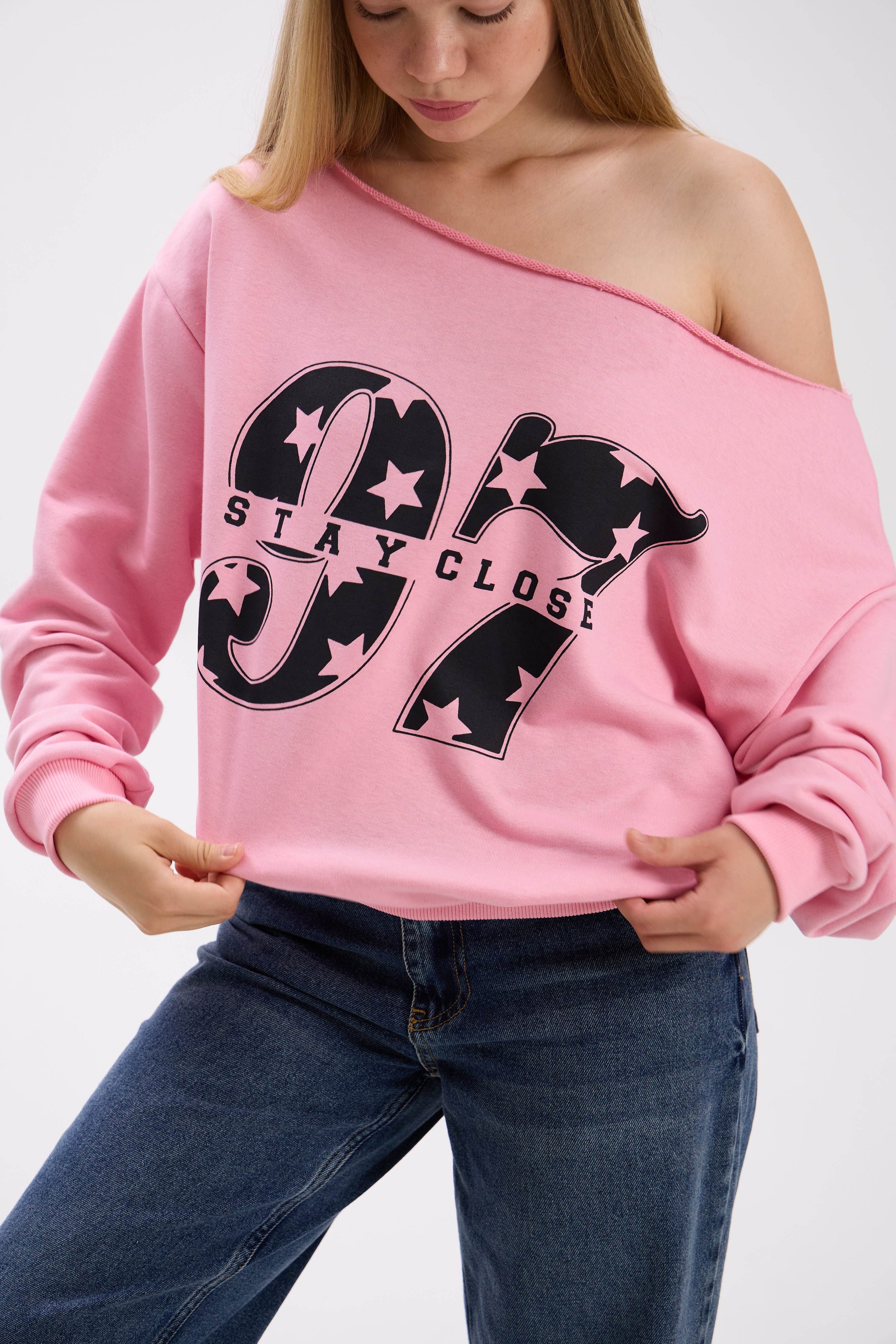 Close-up of pink off-shoulder sweatshirt with large black “Stay Close 97” print, styled with blue jeans.