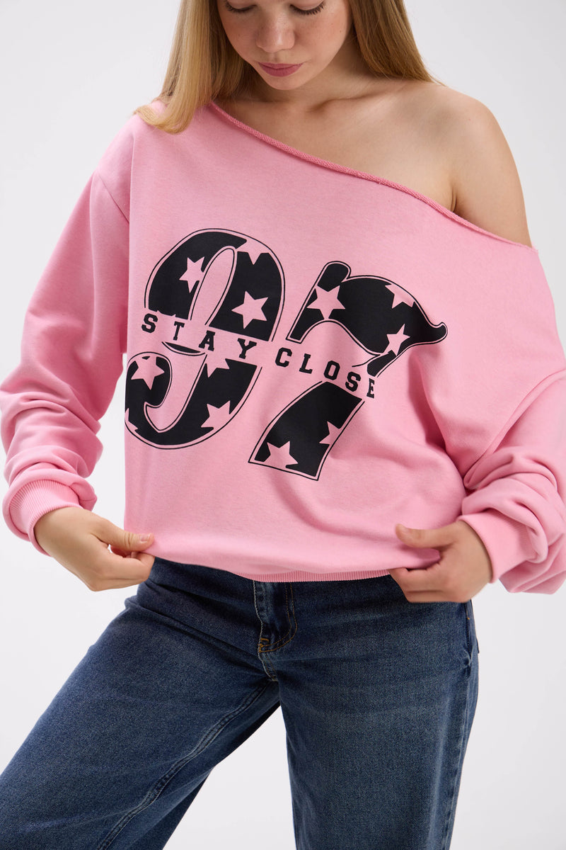 Close-up of pink off-shoulder sweatshirt with large black “Stay Close 97” print, styled with blue jeans.