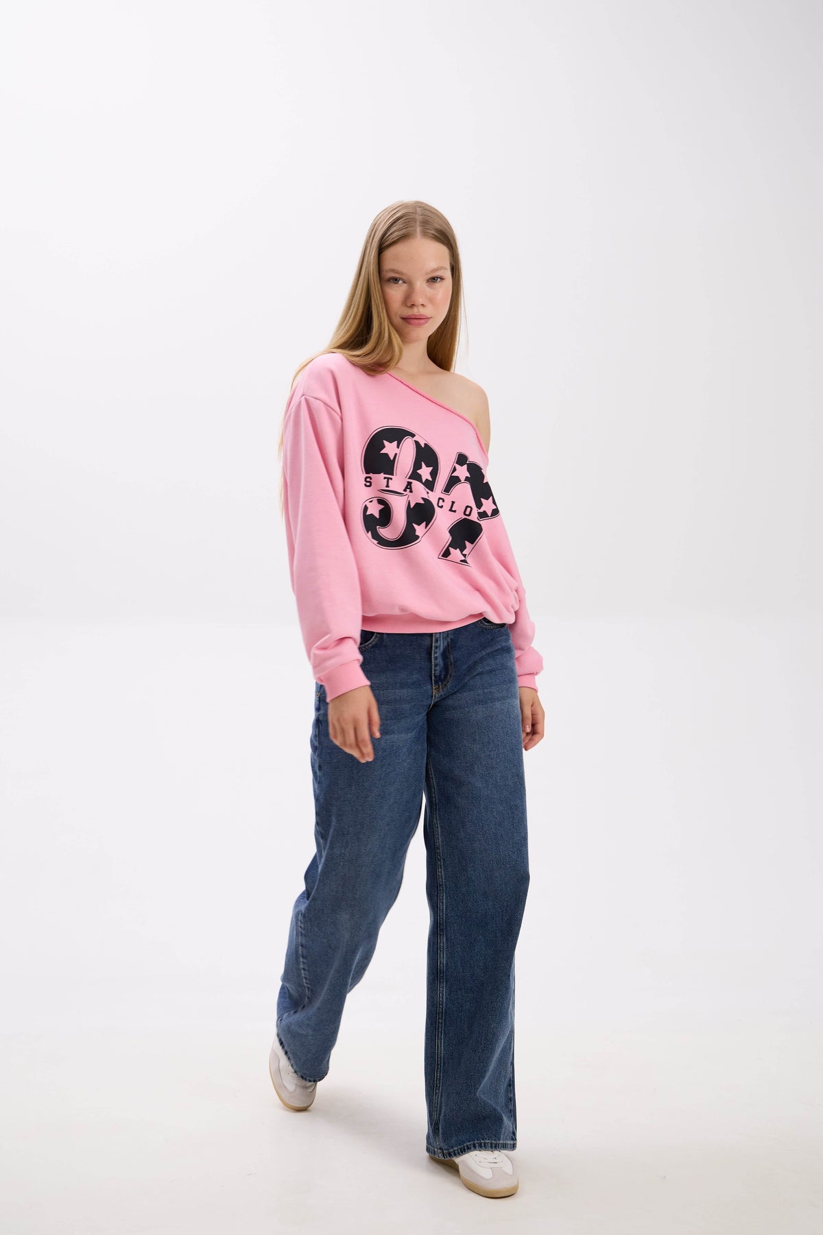 Model walking in a pink off-shoulder sweatshirt with bold black “Stay Close 97” print, styled with blue jeans and sneakers.