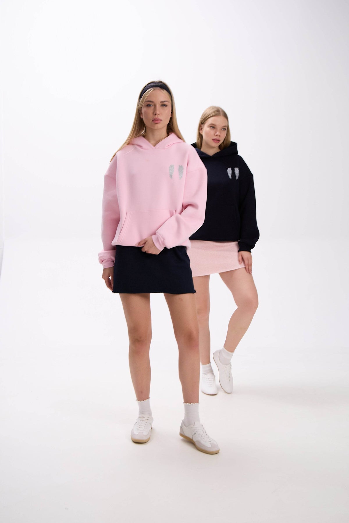 Two female models wearing wing-detail oversized hoodies. The model in front is dressed in a pastel pink hoodie with a navy mini skirt, while the model behind is styled in a navy hoodie with a pale pink mini skirt. Both wear white sneakers, posing in a clean studio background.