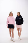 Two women wearing pastel hoodies with angel wing embroidery, one in pink with a navy skirt and the other in black with a light pink skirt, both styled with white sneakers on a clean white background.