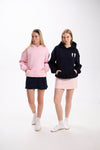 Two women modeling casual hoodies with angel wing embroidery, one in a soft pink hoodie with a navy skirt and the other in a black hoodie with a light pink skirt, both posing with hands in pockets against a white background.