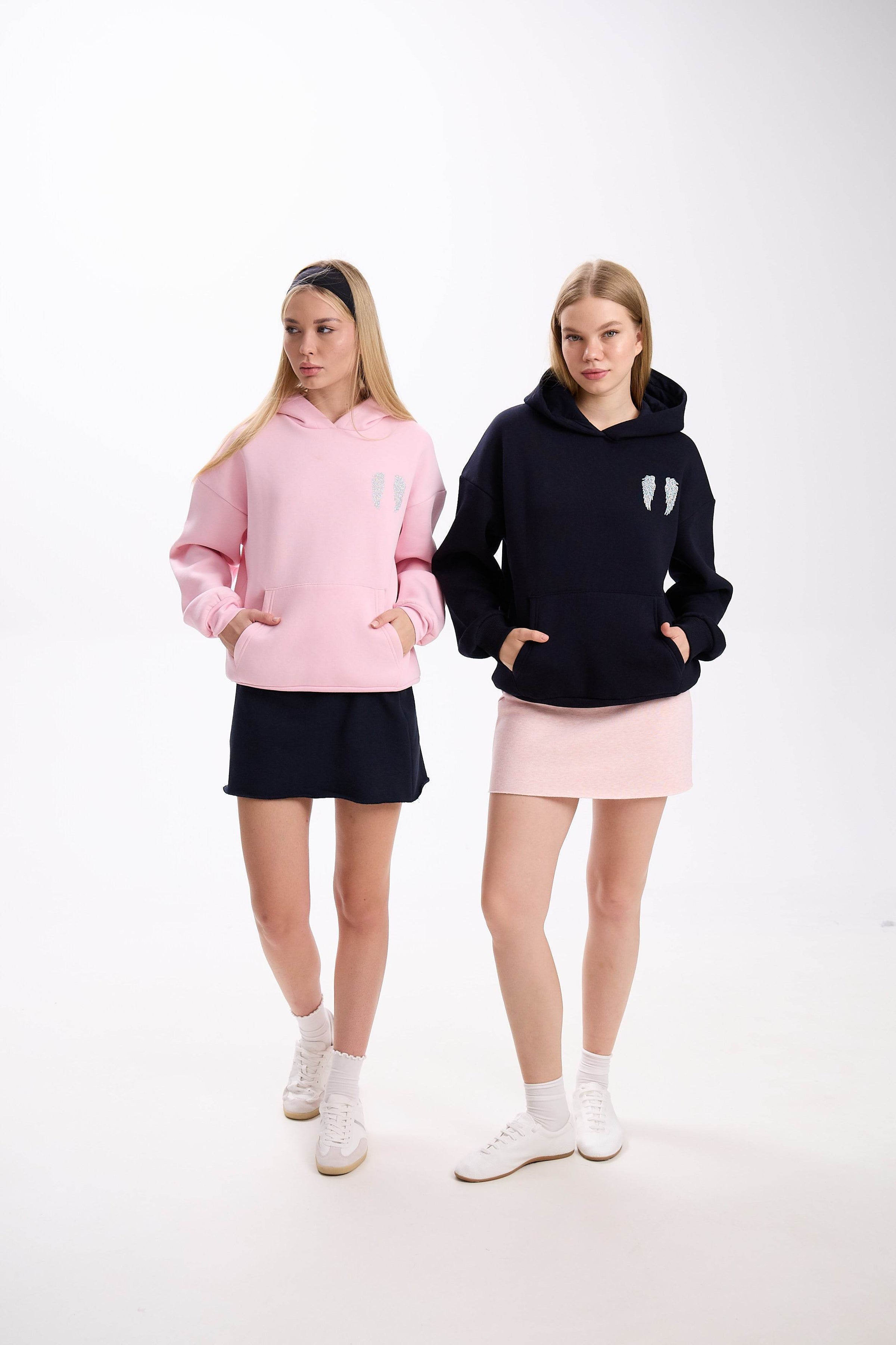 Two women modeling casual hoodies with angel wing embroidery, one in a soft pink hoodie with a navy skirt and the other in a black hoodie with a light pink skirt, both posing with hands in pockets against a white background.