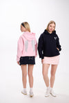 Two women showcasing casual hoodies with angel wing embroidery, one in a pink hoodie with a navy skirt seen from the back, and the other in a black hoodie with a light pink skirt posing from the front, both styled with white sneakers on a white background.