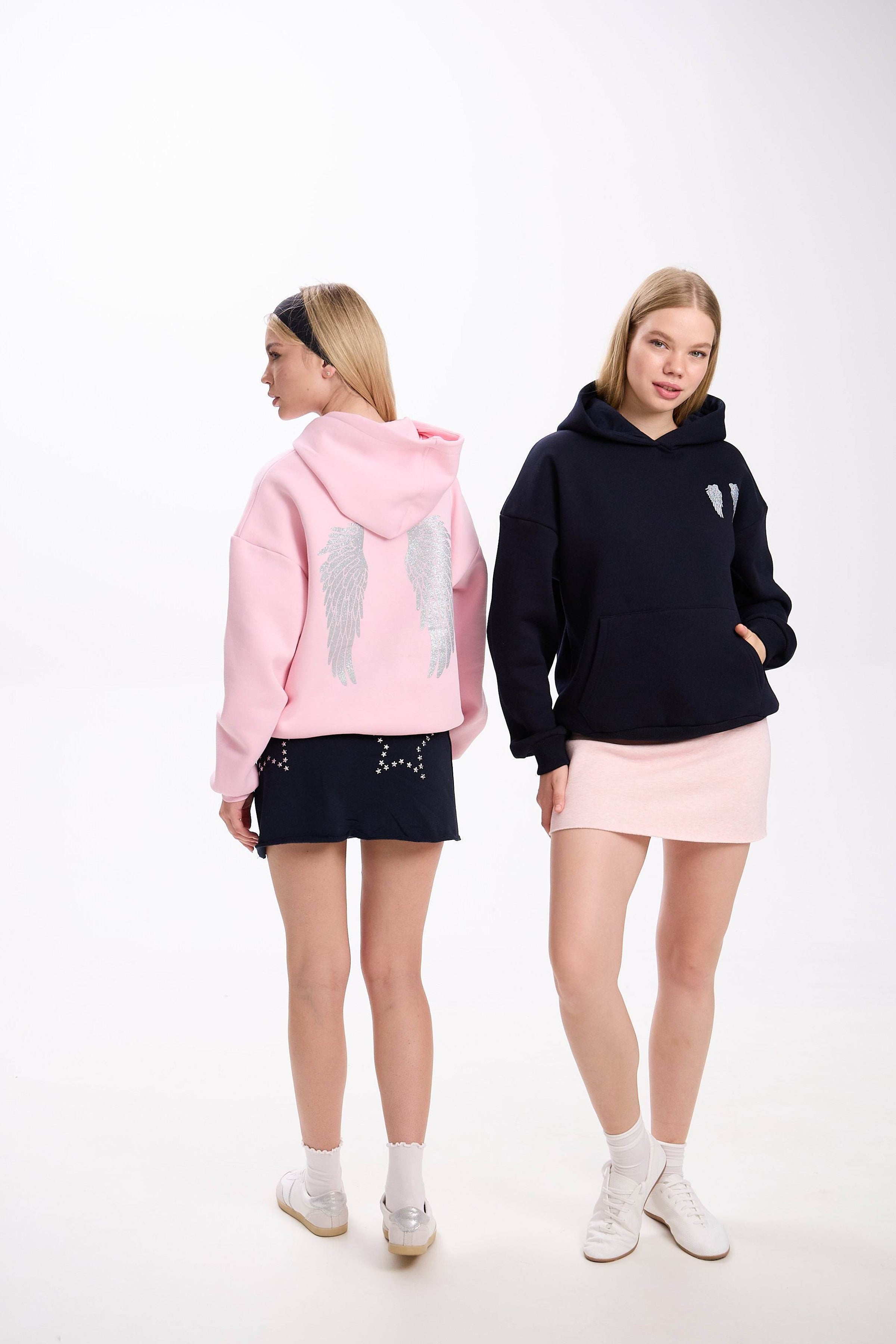 Two women showcasing casual hoodies with angel wing embroidery, one in a pink hoodie with a navy skirt seen from the back, and the other in a black hoodie with a light pink skirt posing from the front, both styled with white sneakers on a white background.