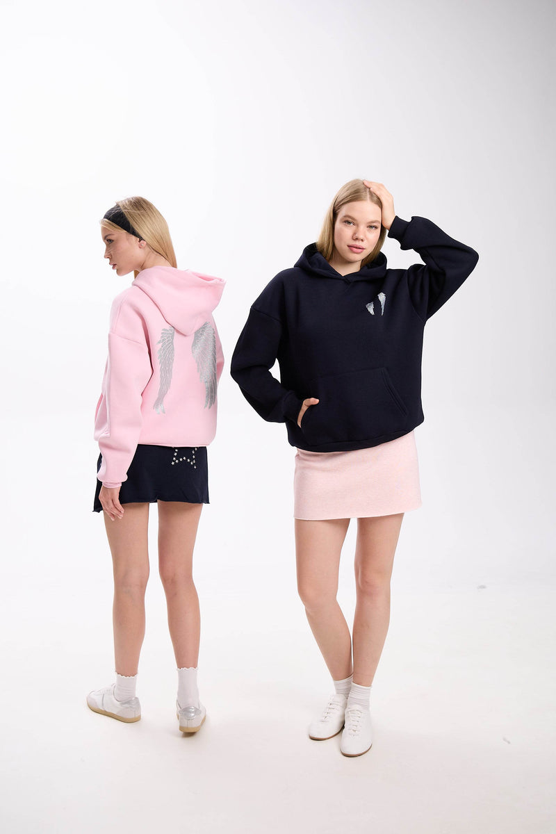 Two women in casual hoodies with angel wing embroidery, one wearing a pink hoodie with navy skirt shown from the side, and the other in a black hoodie with light pink skirt posing with her hand on her head, styled with white sneakers on a plain white background.