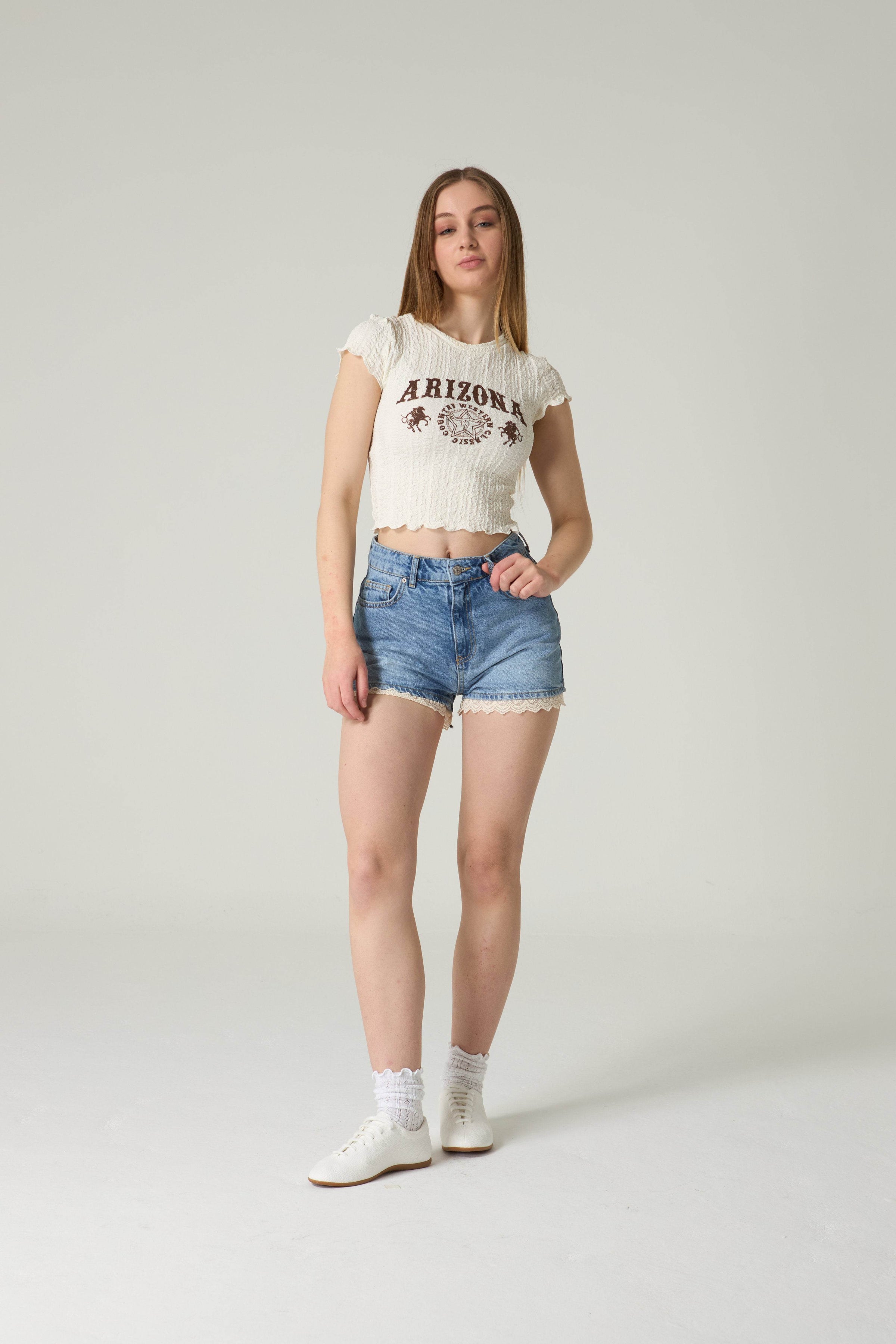 Woman wearing a white crop top with text and denim shorts on a plain background.