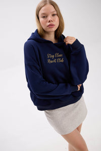 Person wearing a navy hoodie with "Stay Close Resort Club" text and white skirt, standing against a white background
