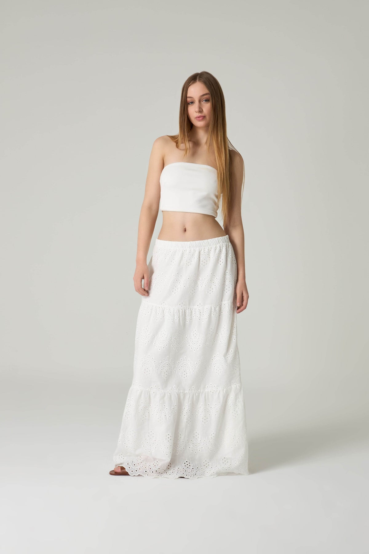 Woman wearing a white strapless top and long white skirt on a plain background.