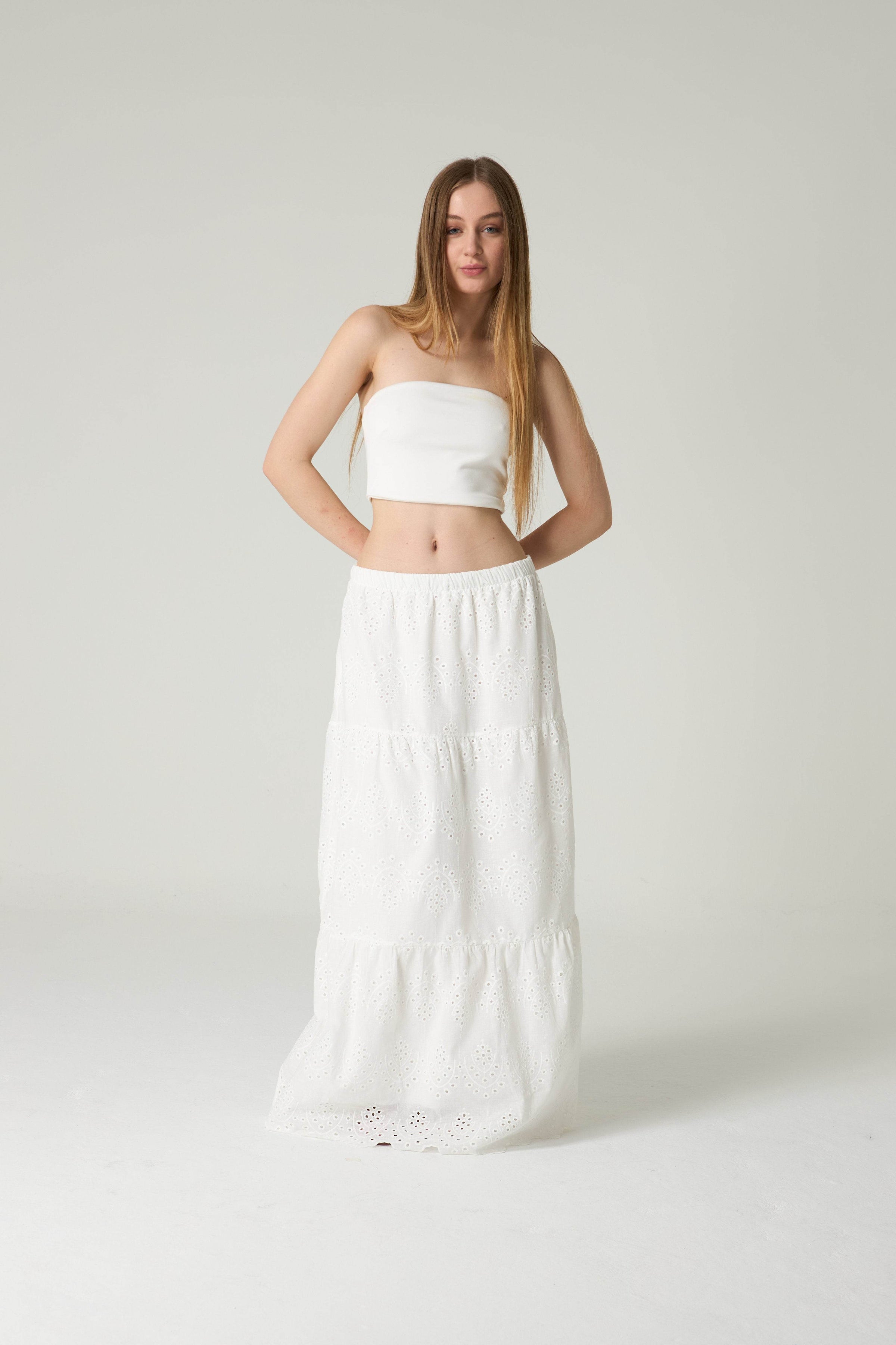 Woman wearing a white strapless top and long white skirt on a light gray background.