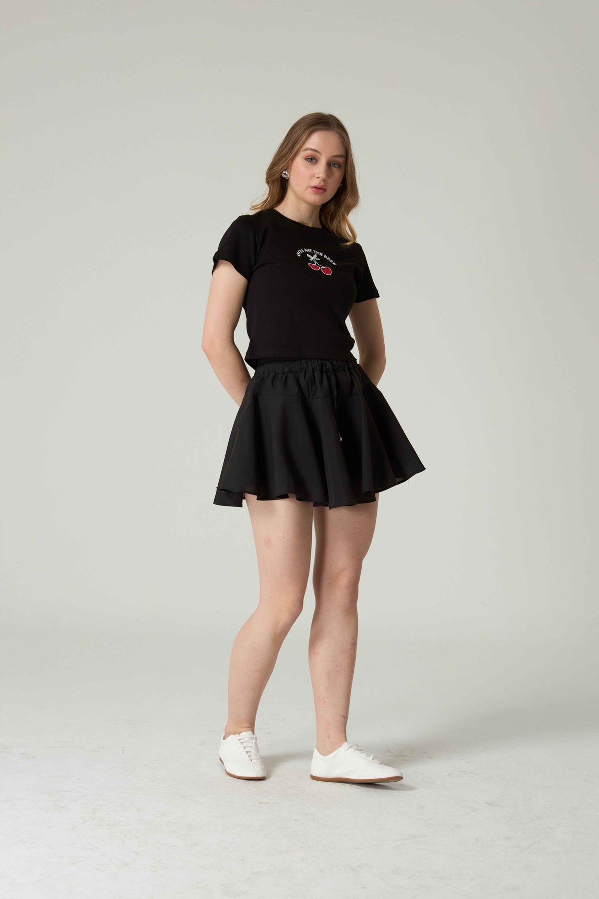 Woman wearing a black t-shirt with cherry embroidery and skirt on a plain background.