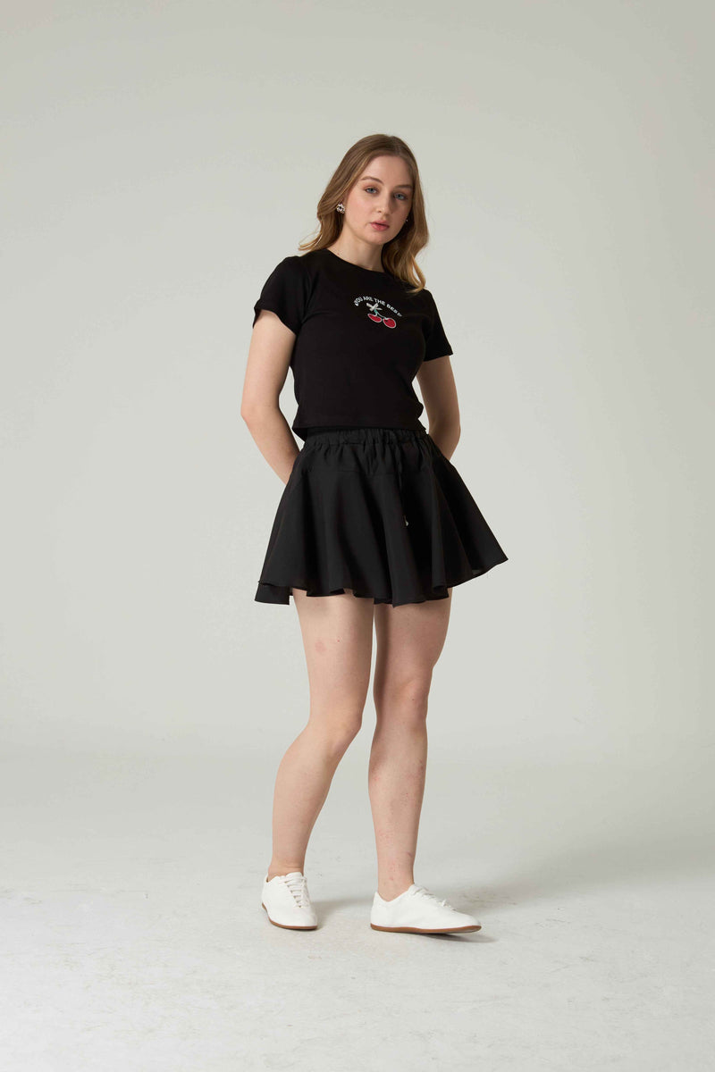 Woman wearing a black t-shirt with cherry embroidery and skirt on a plain background.