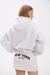 Model in a white hoodie and mini skirt with bold “New York City” text on the back.