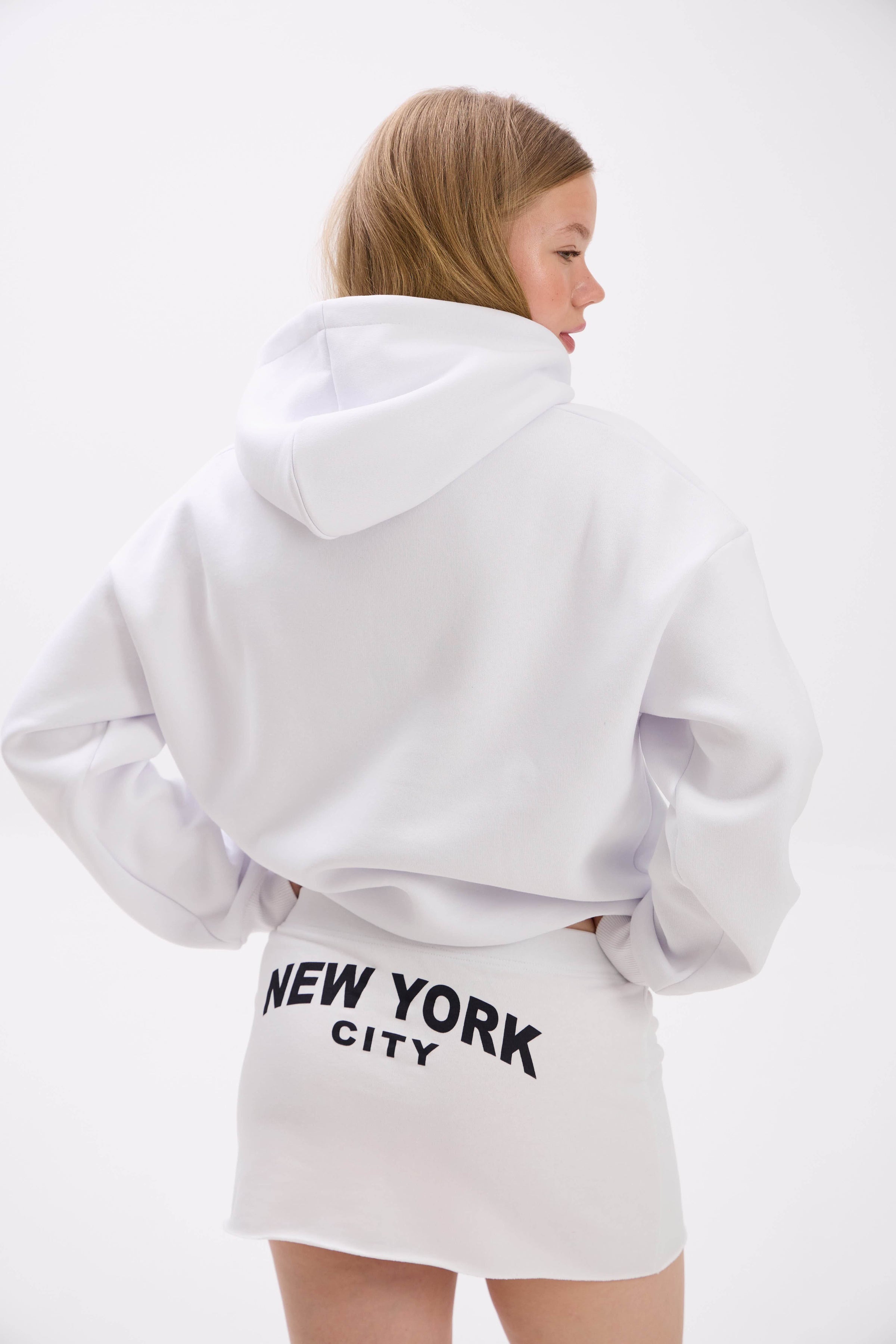 Model in a white hoodie and mini skirt with bold “New York City” text on the back.
