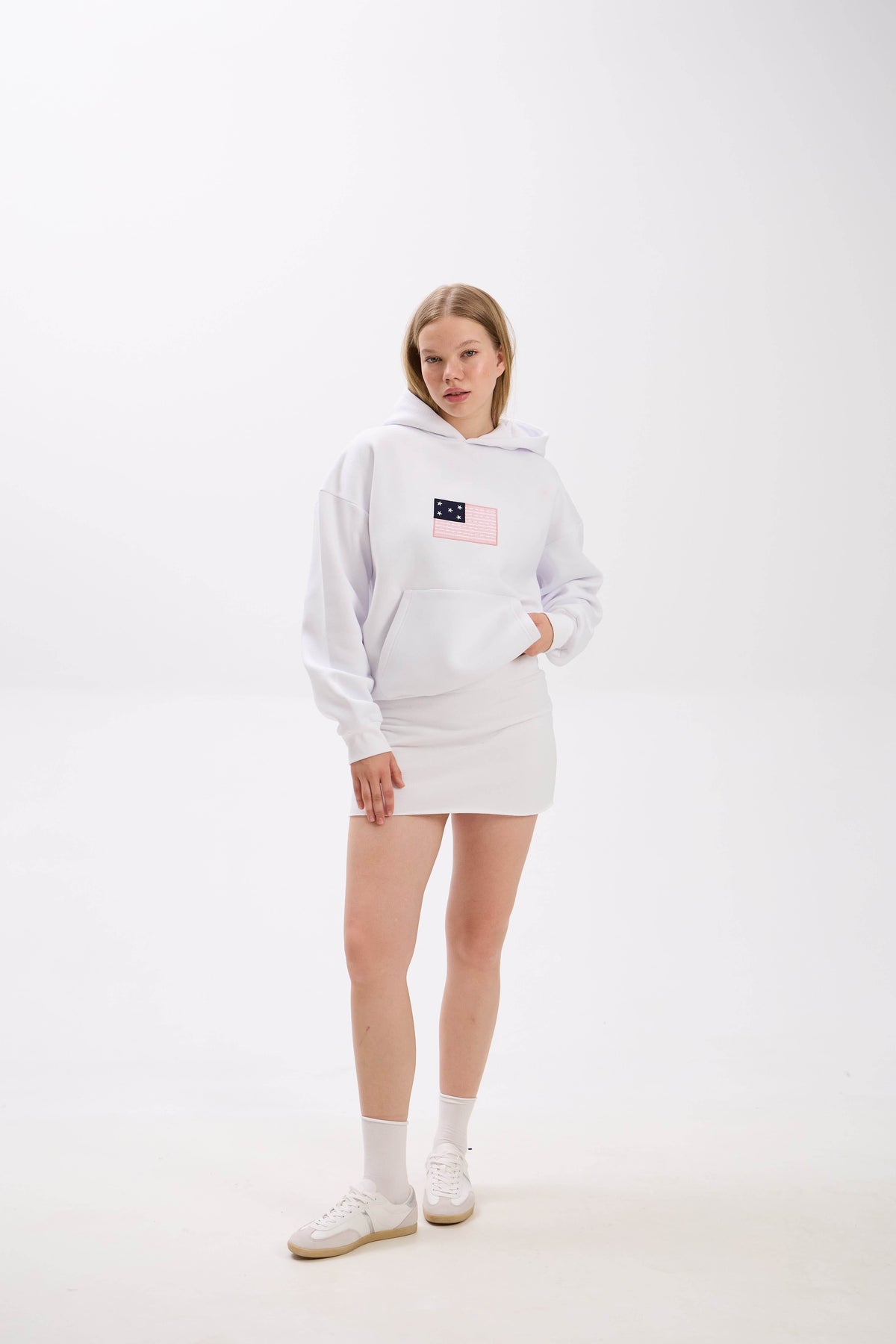 Model wearing a white hoodie with front patch detail, paired with a white mini skirt and white sneakers.