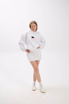 Model in a white hoodie with front patch detail, styled with a white mini skirt, socks, and sneakers.