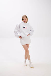 Model in a white hoodie with front patch detail, styled with a white mini skirt, socks, and sneakers.
