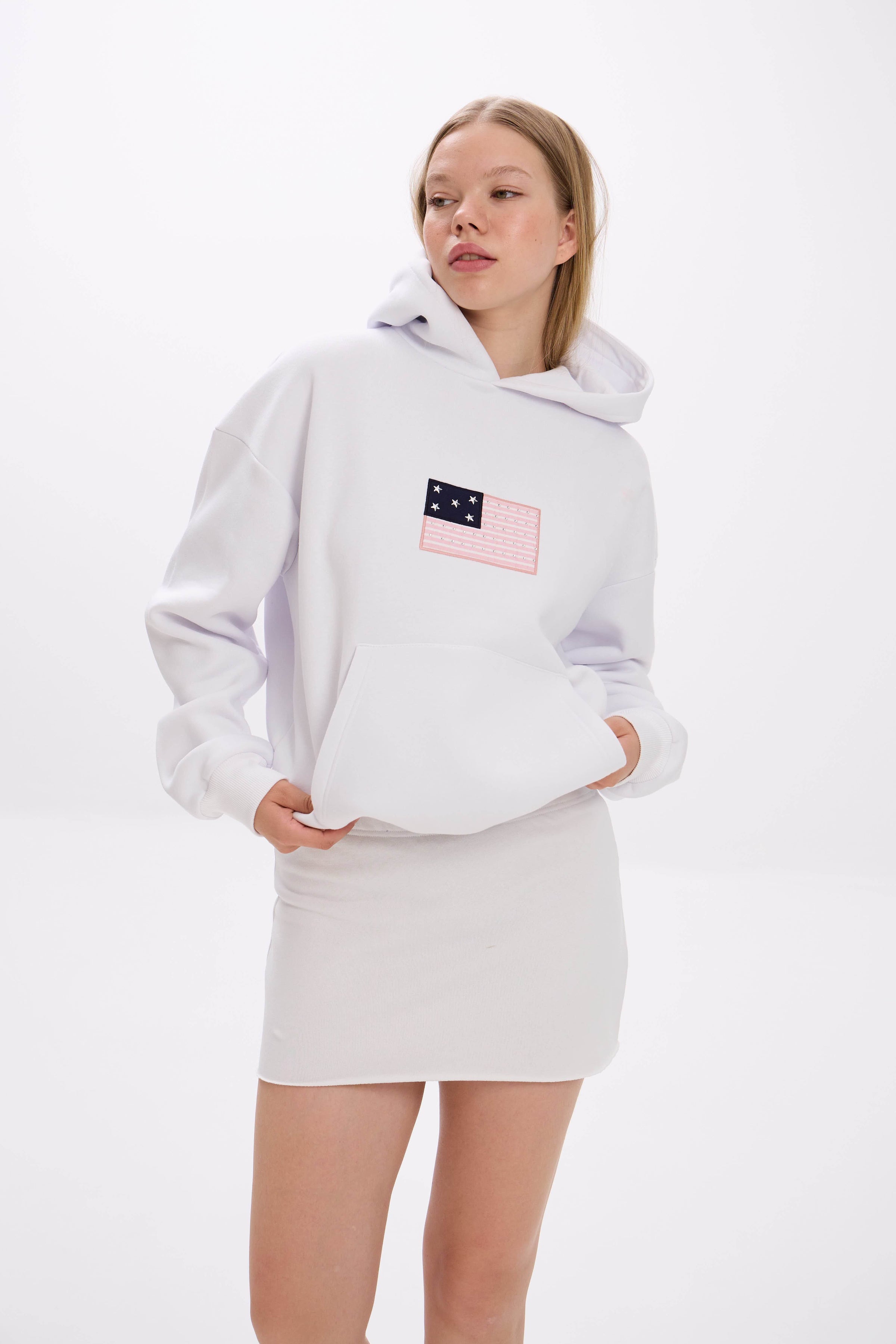 Model wearing a white hoodie with front patch detail, styled with a white mini skirt.