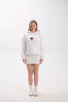 Model in a white hoodie with front patch detail, paired with a white mini skirt, socks, and sneakers.