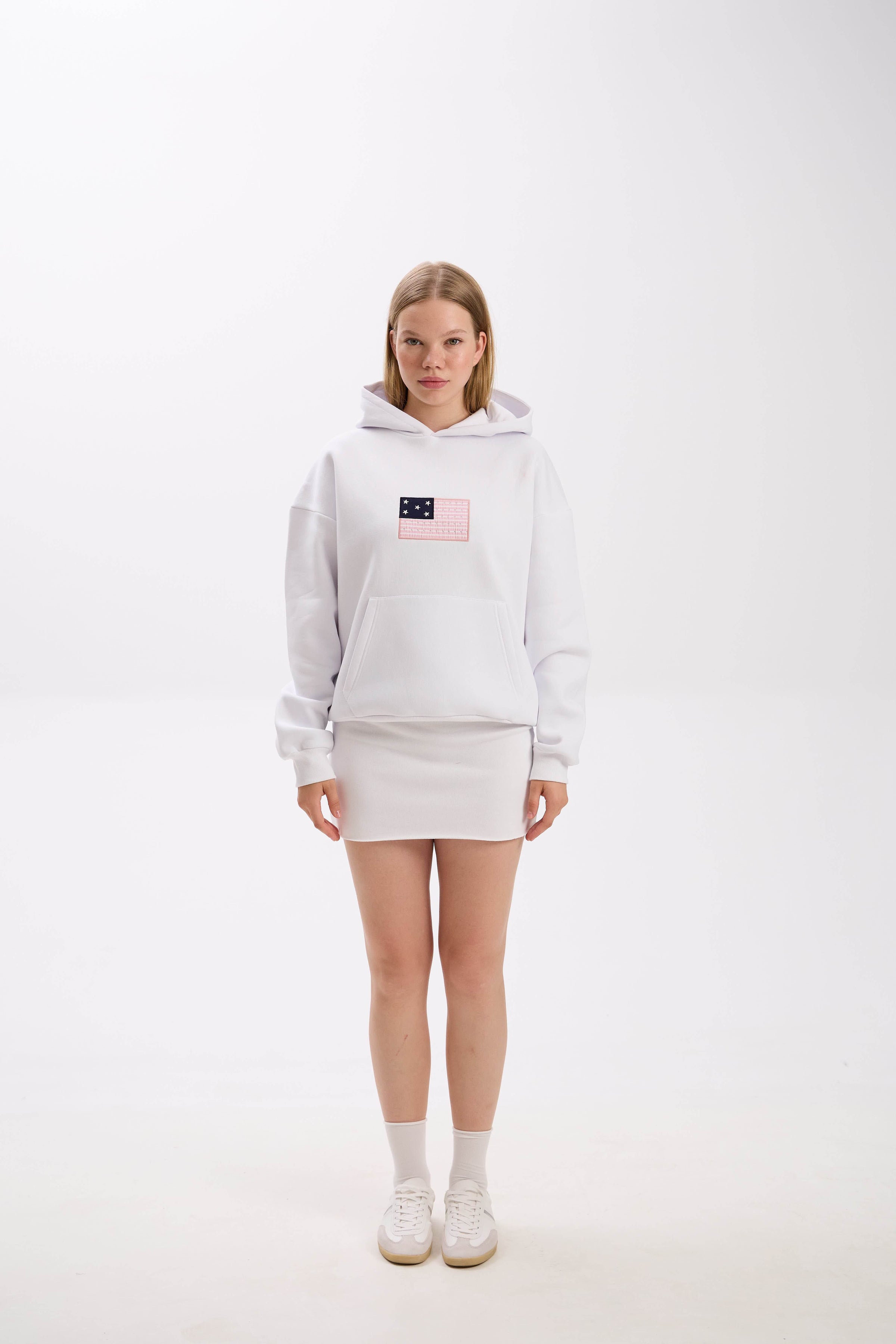Model in a white hoodie with front patch detail, paired with a white mini skirt, socks, and sneakers.