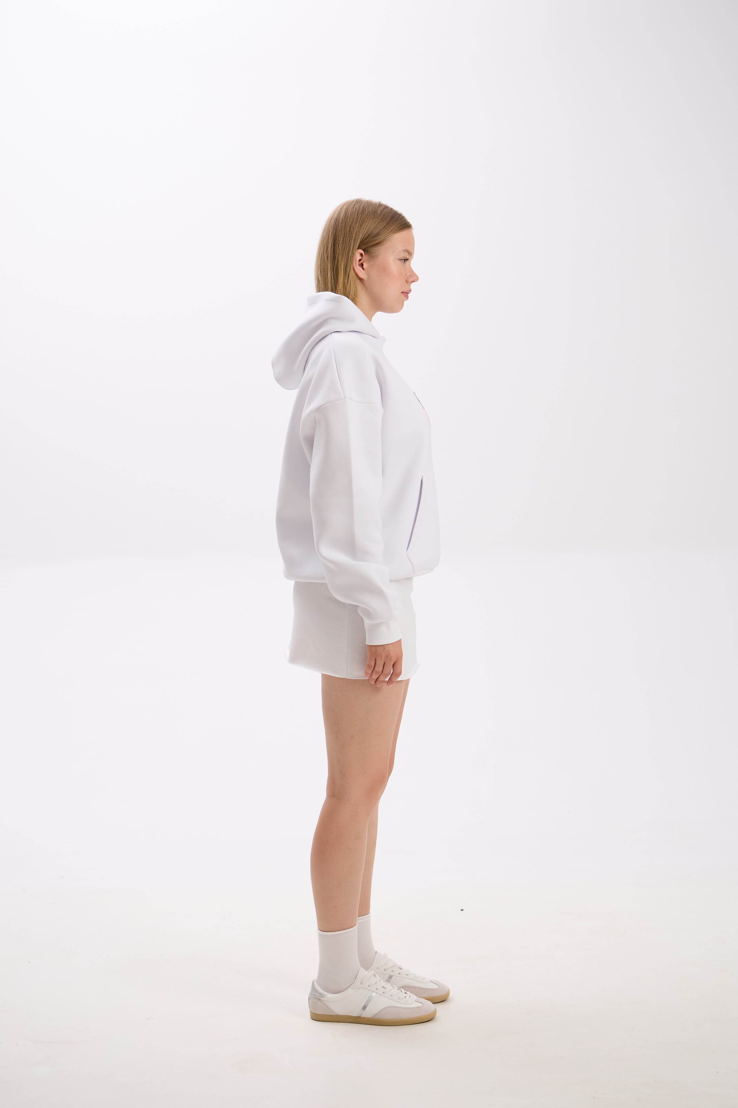 Side profile of model in a white hoodie paired with a white mini skirt, socks, and sneakers.
