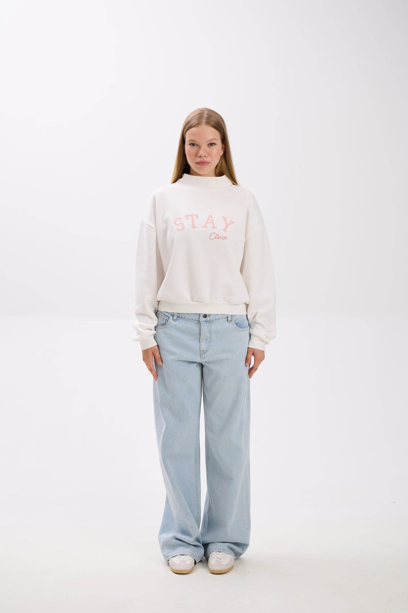 Person wearing a white sweatshirt with text and light blue jeans on a white background