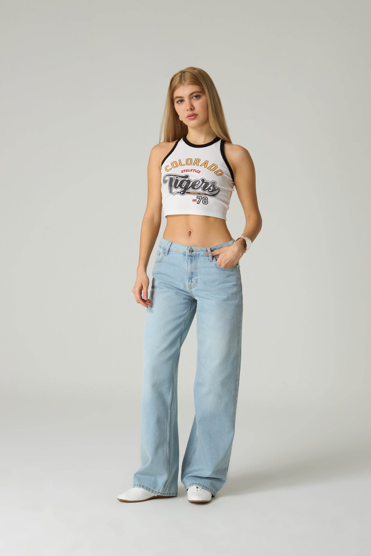 Woman wearing a white tank top with 'Colorado Tigers' text and light blue jeans on a plain background.