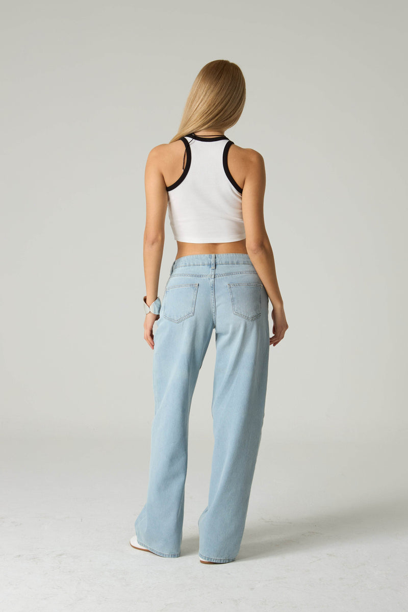 Woman wearing a white crop top with black straps and light blue jeans on a plain background.