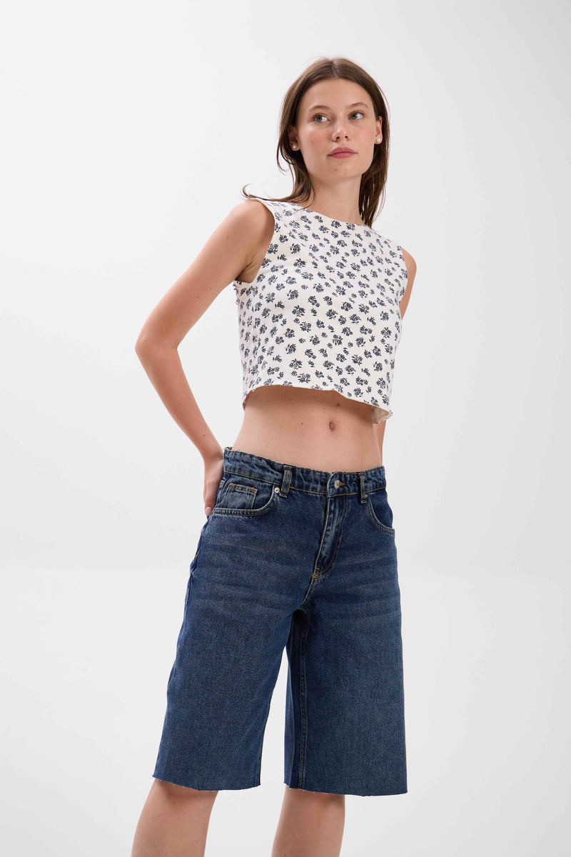 Woman wearing a white sleeveless top with black patterns and blue denim shorts on a white background.