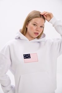 Close-up of a woman wearing a white hoodie with a pink and navy American flag-inspired patch featuring stars and stripes on the chest, adjusting her hair with one hand, against a plain background