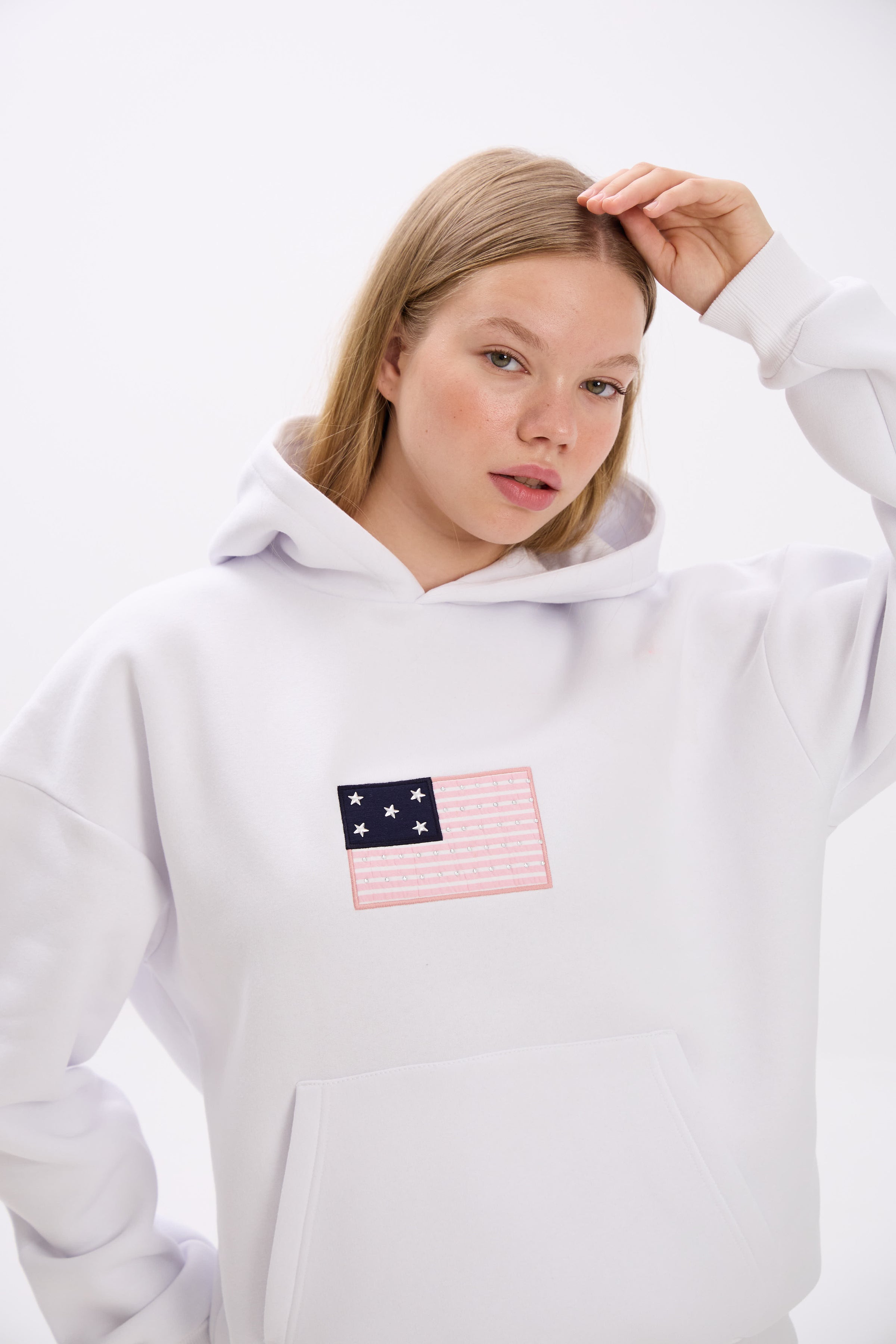 Close-up of a woman wearing a white hoodie with a pink and navy American flag-inspired patch featuring stars and stripes on the chest, adjusting her hair with one hand, against a plain background