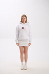 Full-body photo of a woman wearing a white hoodie with a pink and navy American flag-inspired patch on the chest, paired with a matching white mini skirt, white socks, and white sneakers, standing against a plain studio background.