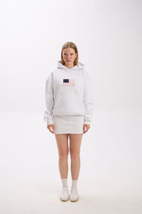 Full-body photo of a woman wearing a white hoodie with a pink and navy American flag-inspired patch on the chest, paired with a matching white mini skirt, white socks, and white sneakers, standing against a plain studio background.