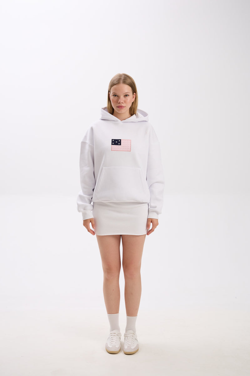 Full-body photo of a woman wearing a white hoodie with a pink and navy American flag-inspired patch on the chest, paired with a matching white mini skirt, white socks, and white sneakers, standing against a plain studio background.