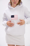 Close-up of a woman wearing a white hoodie with a pink and navy American flag-inspired patch on the chest, styled with a matching white mini skirt, showing hands resting on the hoodie pocket against a plain background.
