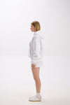 Side profile view of a woman wearing a white hoodie with a front pocket and a small American flag-inspired patch, styled with a matching white mini skirt and white sneakers, standing against a plain background.