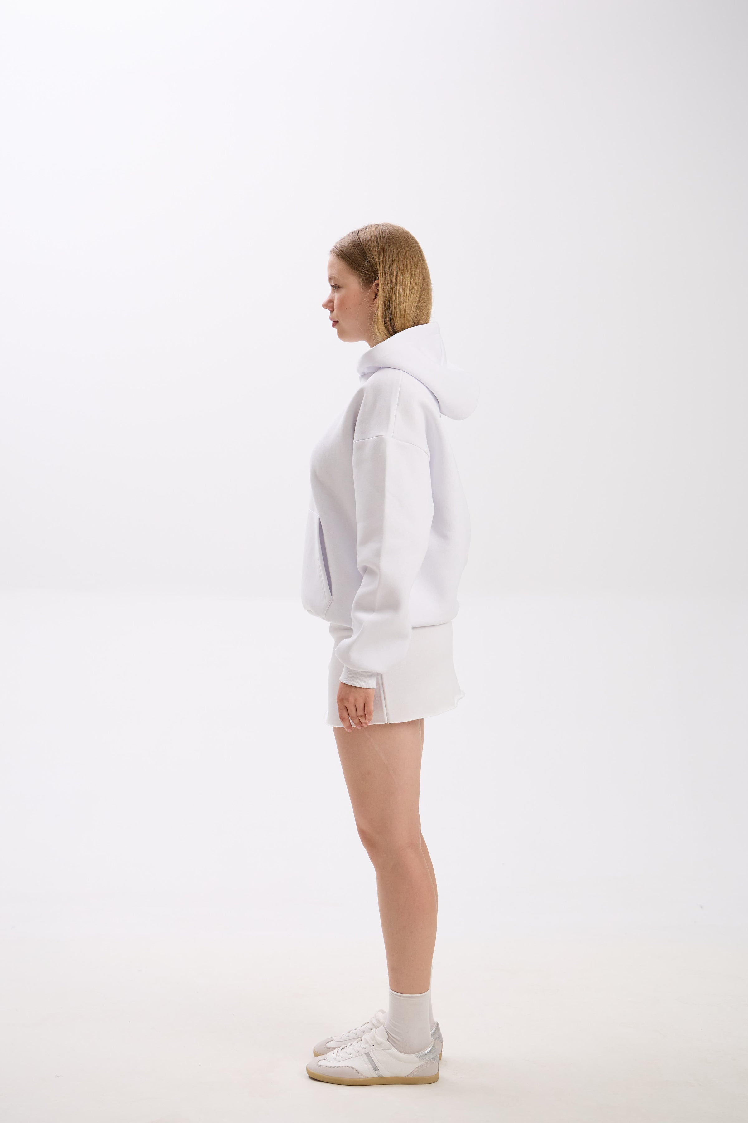 Side profile view of a woman wearing a white hoodie with a front pocket and a small American flag-inspired patch, styled with a matching white mini skirt and white sneakers, standing against a plain background.