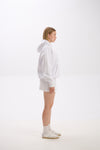 Side profile view of a woman wearing a white hoodie with a front pocket and hood, styled with a matching white mini skirt, white socks, and white sneakers, standing against a plain background.