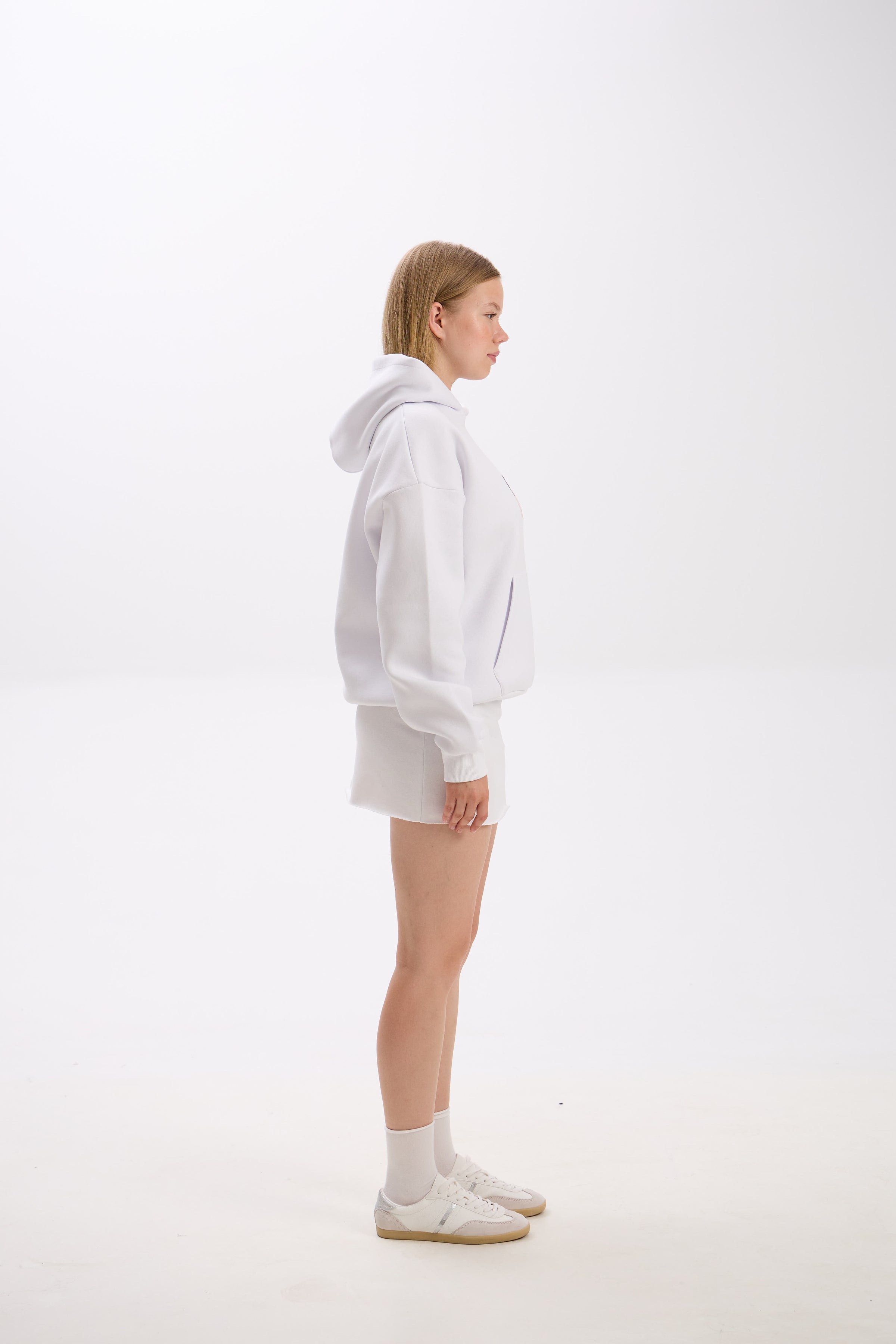 Side profile view of a woman wearing a white hoodie with a front pocket and hood, styled with a matching white mini skirt, white socks, and white sneakers, standing against a plain background.