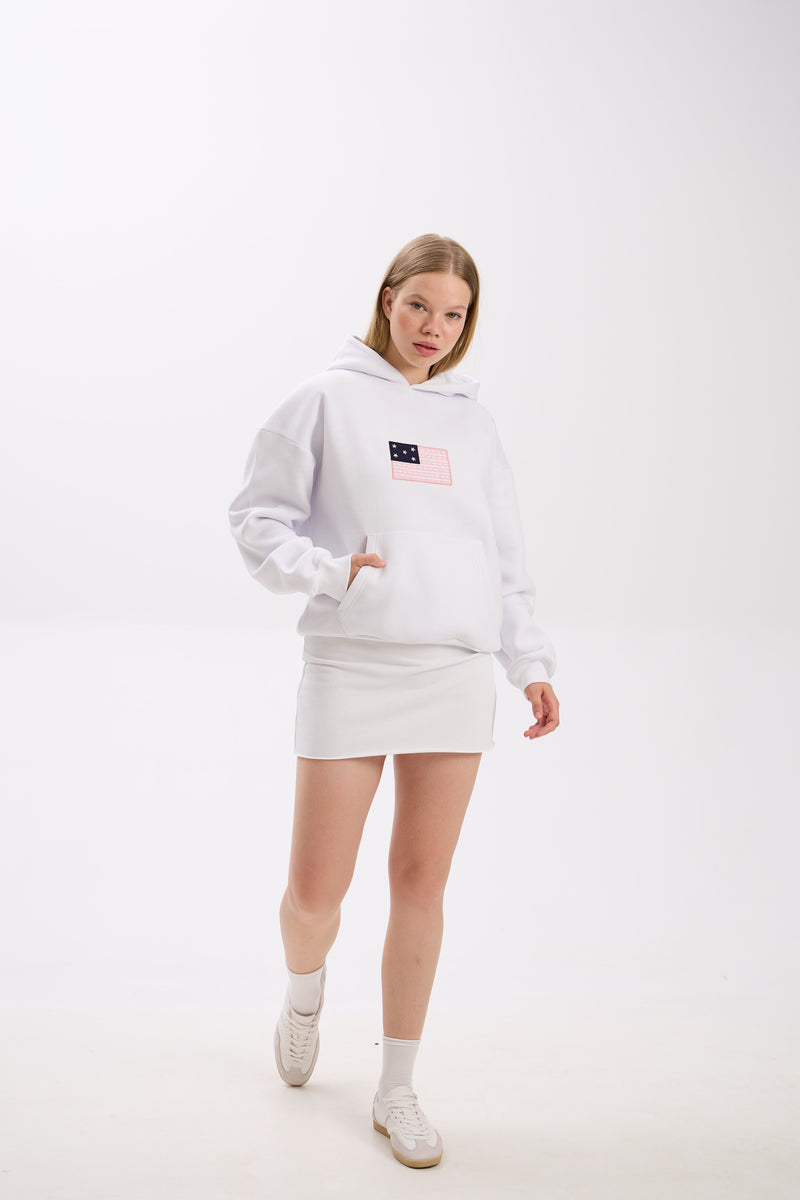 Woman wearing a white hoodie with a pink and navy flag patch detail on the chest, styled with a matching white mini skirt and white sneakers, posing with one hand in the hoodie pocket while walking forward against a plain background.