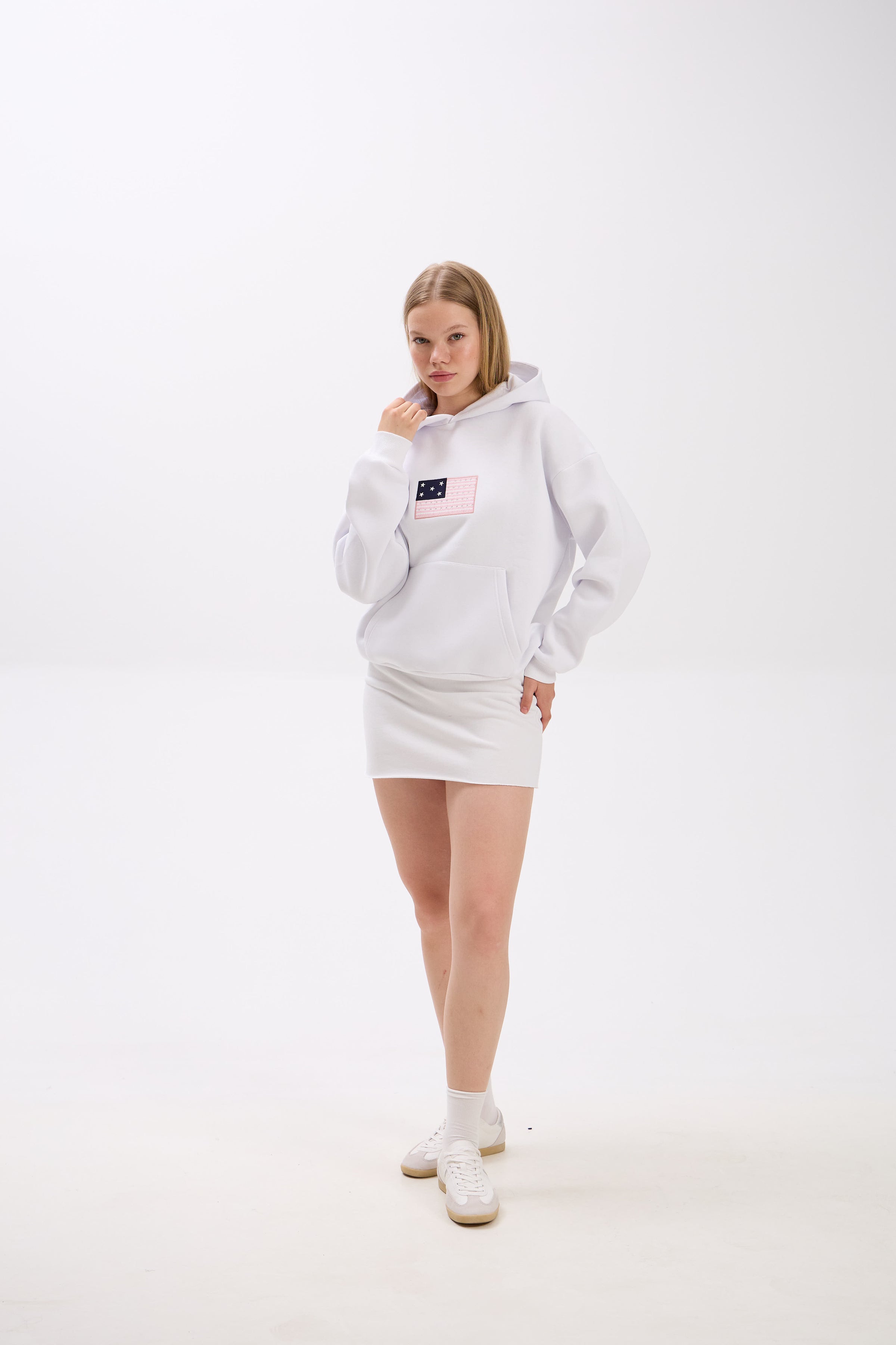 Woman posing in a white hoodie featuring a pink and navy flag patch detail on the chest, paired with a white mini skirt and white sneakers, standing against a plain white background with one hand slightly lifting the hoodie collar.